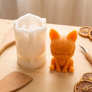 May include: A translucent white silicone mould shaped like a cat and a finished orange wax figure of a cat with pointed ears and a stern facial expression. The mould is cylindrical with a hollow centre, and the wax figure sits upright on a surface.