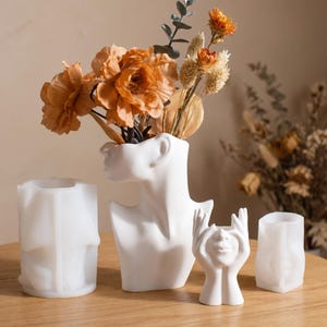 May include: A collection of four white silicone molds for crafting, including a large bust-shaped vase mold, a smaller mold featuring hands covering a face, and two cylindrical molds with embossed human torso designs, all arranged on a wooden surface.