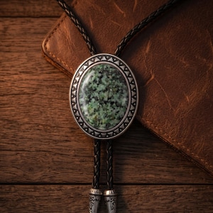 Pode incluir: Uma gravata bolo com uma peça central oval com incrustação de pedra salpicada verde e preta. A moldura de metal tem um padrão triangular decorativo ao longo da borda. Inclui um cordão de couro trançado preto com pontas de metal prateado.