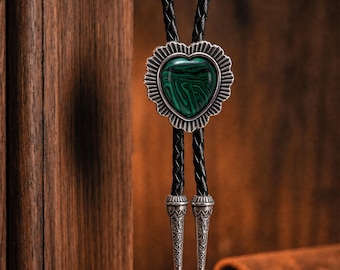 May include: A bolo tie featuring a heart-shaped green malachite stone set in a scalloped silver-tone metal frame. The pendant is attached to a black braided leather cord that ends in two ornate, tapered silver-tone metal tips with engraved details.