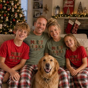 May include: A family of four wearing matching t-shirts with the text Merry Christmas and plaid pajama pants. The two adults wear olive green shirts, while the two children wear red shirts. A golden retriever sits in the center of the group.