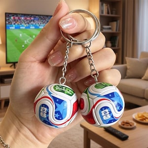 May include: A double keychain featuring two miniature soccer balls with white, blue, red, and green panels. Each ball displays a blue star pattern and a small rectangular logo with the text FIFA 26. The balls are attached to silver metal chains.