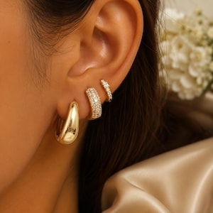 May include: A set of three gold-toned earrings worn in a single ear. The largest is a smooth, polished gold hoop. The middle earring is a thicker gold hoop encrusted with small clear stones. The smallest earring is a thin gold hoop with stones.