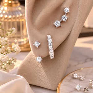 May include: A set of silver-toned earrings featuring a three-stone ear climber, a small hoop earring encrusted with clear stones, and two single-stone stud earrings. The jewelry pieces are displayed on a beige ear-shaped stand with additional studs.