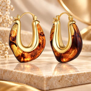 May include: A pair of hoop earrings featuring a polished gold-tone metal frame integrated with a translucent, amber-colored resin element that displays a swirling, marbled pattern throughout the material, complete with a secure hinged clasp.