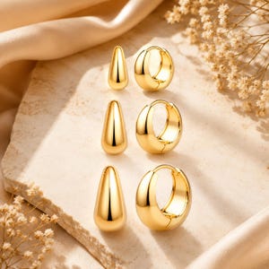 May include: A set of three pairs of gold-toned earrings arranged in a vertical column. Each pair consists of one smooth, teardrop-shaped stud earring and one thick, polished hoop earring. The jewelry features a reflective, high-shine metallic finish.