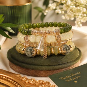 May include: A stack of four beaded bracelets featuring olive green, cream, and marbled gray beads with gold-toned metal spacers and accents. A central gold-toned charm with a dangling clear triangular pendant sits at the front of the bracelet stack.