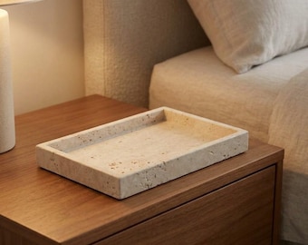 May include: A rectangular tray crafted from beige travertine stone, featuring a porous, textured surface with natural pitting and variations in color. The tray has a low, solid rim and a flat base, showcasing the organic, earthy aesthetic of the stone.