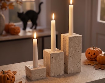 May include: Three beige travertine stone candle holders of varying heights, each featuring a square base and a single lit white taper candle. The textured, porous stone surfaces are arranged in a row, creating a tiered display of light.
