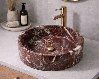 May include: A round vessel sink made of deep red marble with prominent white veining, featuring a polished finish. The sink is equipped with a central gold-toned drain plug and is paired with a matching gold-toned faucet mounted on the countertop.