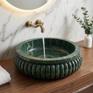 May include: A round, dark green marble vessel sink with a fluted exterior design. A brass wall-mounted faucet is positioned above the basin, with a steady stream of water flowing into the center of the bowl against a white marble wall.