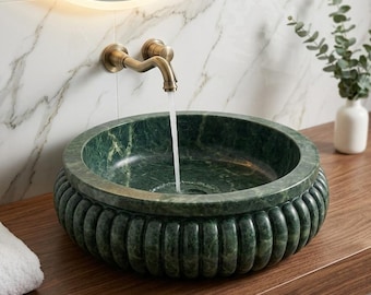 May include: A round, dark green marble vessel sink with a fluted exterior design. A brass wall-mounted faucet is positioned above the basin, with a steady stream of water flowing into the center of the bowl against a white marble wall.