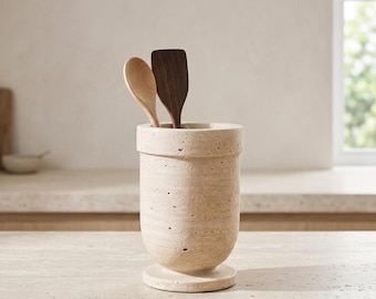 May include: A cylindrical beige travertine utensil holder with a porous, natural stone texture and a wide circular base. Two wooden kitchen spoons, one light and one dark, are placed inside the holder, extending from the top opening.