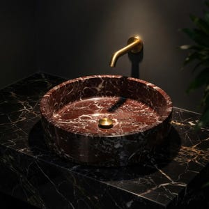 May include: A circular deep red marble vessel sink with white veining, featuring a matching red marble countertop with white and gold veining. A wall-mounted brushed gold faucet and a matching circular gold drain plug are installed above the sink.