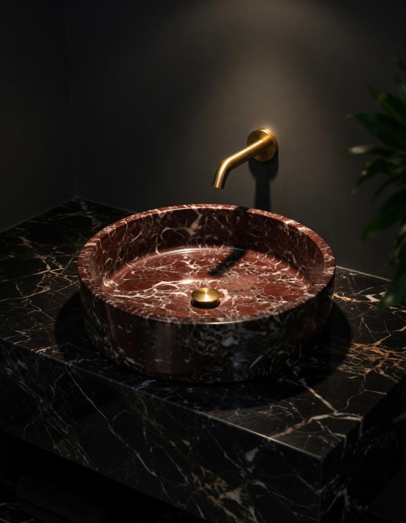 May include: A circular deep red marble vessel sink with white veining, featuring a matching red marble countertop with white and gold veining. A wall-mounted brushed gold faucet and a matching circular gold drain plug are installed above the sink.