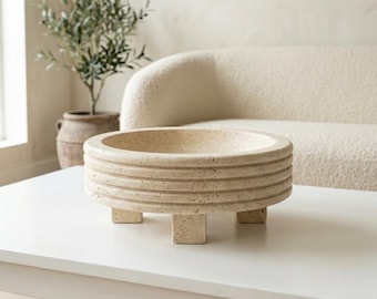 May include: A circular beige travertine bowl with a porous, speckled texture. The exterior features three horizontal ridges encircling the body, and the vessel is supported by three short, rectangular feet extending from the base.