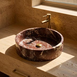 May include: A circular vessel sink crafted from deep red marble with prominent white veining. The sink features a central brass drain plug and is paired with a matching brass faucet, both set against a light beige stone countertop and backsplash.