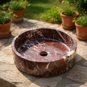 May include: A circular vessel sink crafted from deep red marble with prominent white veining throughout the stone. The basin features a smooth, polished finish and a centered circular drain hole, showcasing the natural patterns of the marble.