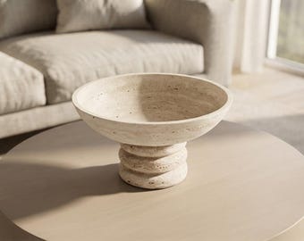 May include: A beige travertine bowl with a wide, circular basin and a thick, tiered pedestal base. The stone features a natural, porous texture with visible pits and variations in color throughout the surface of the decorative vessel.