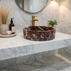 May include: A circular vessel sink crafted from deep red marble with prominent white veining, positioned on a white marble countertop. The sink features a brushed gold faucet, set against a white marble wall with a round mirror and gold lighting.