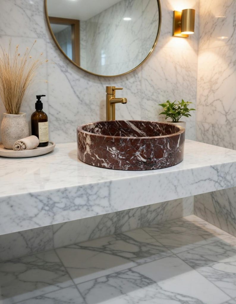 May include: A circular vessel sink crafted from deep red marble with prominent white veining, positioned on a white marble countertop. The sink features a brushed gold faucet, set against a white marble wall with a round mirror and gold lighting.
