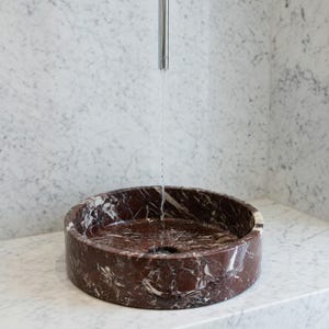 May include: A circular, deep red marble vessel sink with prominent white veining. A thin, vertical stream of water flows from a tall, slender, polished chrome faucet centered directly above the basin, which is set into a white marble counter.