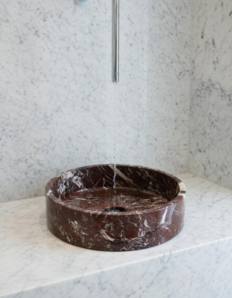 May include: A circular, deep red marble vessel sink with prominent white veining. A thin, vertical stream of water flows from a tall, slender, polished chrome faucet centered directly above the basin, which is set into a white marble counter.