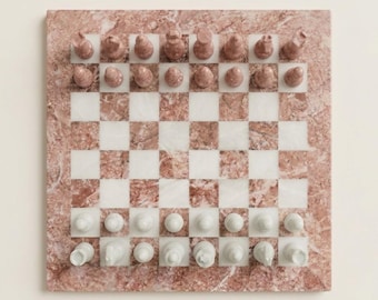 May include: A square chess set featuring a board with alternating rose-pink and white marble squares. The set includes sixteen rose-pink marble chess pieces and sixteen white marble chess pieces arranged in their starting positions on the board.