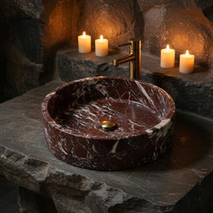 May include: A round vessel sink crafted from deep red marble with prominent white veining. The sink features a polished finish and is equipped with a brass-toned drain plug, paired with a matching brass-toned faucet fixture mounted behind it.