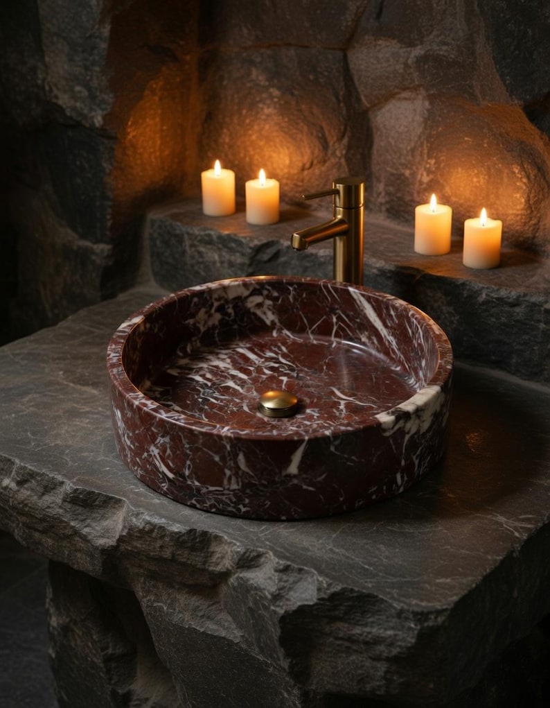 May include: A round vessel sink crafted from deep red marble with prominent white veining. The sink features a polished finish and is equipped with a brass-toned drain plug, paired with a matching brass-toned faucet fixture mounted behind it.