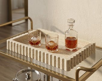 May include: A rectangular beige travertine tray with a scalloped edge design. The tray holds two clear glass tumblers with a diamond-cut pattern and a matching glass decanter, each containing an amber-colored liquid and a spherical ice cube.
