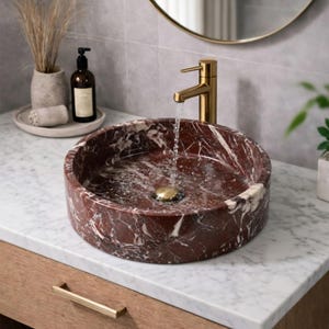 May include: A round, deep red marble vessel sink with white veining, featuring a gold-toned faucet pouring water into the basin. The sink sits on a white marble countertop with a wooden vanity cabinet below, accompanied by a soap dispenser and vase.