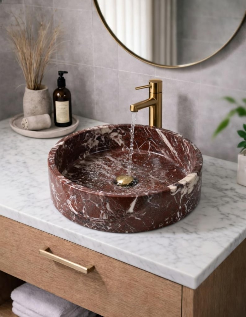 May include: A round, deep red marble vessel sink with white veining, featuring a gold-toned faucet pouring water into the basin. The sink sits on a white marble countertop with a wooden vanity cabinet below, accompanied by a soap dispenser and vase.