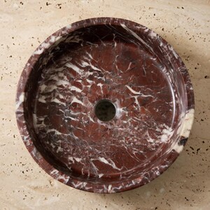 May include: A circular vessel sink crafted from deep red marble with prominent white veining throughout the stone surface. The basin features a smooth, polished interior finish and a centered circular drain opening, showcasing the natural patterns.