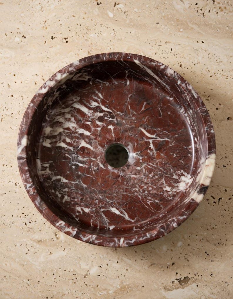May include: A circular vessel sink crafted from deep red marble with prominent white veining throughout the stone surface. The basin features a smooth, polished interior finish and a centered circular drain opening, showcasing the natural patterns.