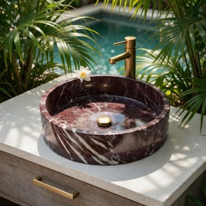 May include: A circular dark red marble vessel sink with white veining, featuring a brushed gold faucet and a matching gold drain plug. The sink is mounted on a light-colored stone countertop with a wooden cabinet base and a gold drawer handle.
