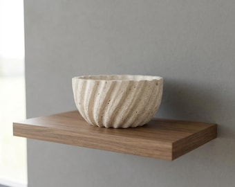 May include: A beige, porous stone bowl with a distinct spiral, fluted texture along its exterior surface. The bowl features a rounded, open top and a textured, natural finish, showcasing the organic, pitted appearance of the stone material.