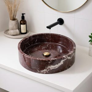 May include: A round vessel sink made of deep red marble with white veining, featuring a polished brass drain plug. A matte black wall-mounted faucet is positioned above the basin, with a thin stream of water flowing into the center of the sink.