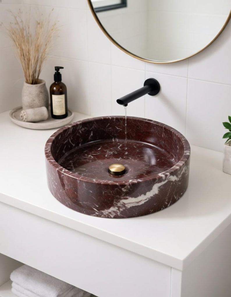 May include: A round vessel sink made of deep red marble with white veining, featuring a polished brass drain plug. A matte black wall-mounted faucet is positioned above the basin, with a thin stream of water flowing into the center of the sink.