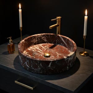 May include: A round, deep red marble vessel sink with white veining, featuring a brushed gold faucet and a matching gold drain plug. The sink is placed on a dark gray countertop, flanked by two tall, thin white candles in gold holders.