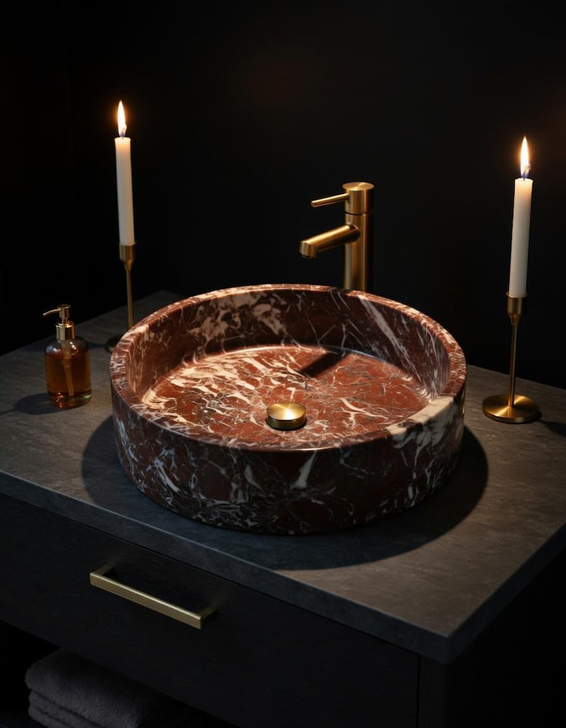 May include: A round, deep red marble vessel sink with white veining, featuring a brushed gold faucet and a matching gold drain plug. The sink is placed on a dark gray countertop, flanked by two tall, thin white candles in gold holders.