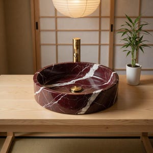 May include: A circular vessel sink crafted from deep red marble with prominent white veining. The basin features a central brass drain plug and is paired with a tall, cylindrical brass faucet fixture centered directly behind the bowl.