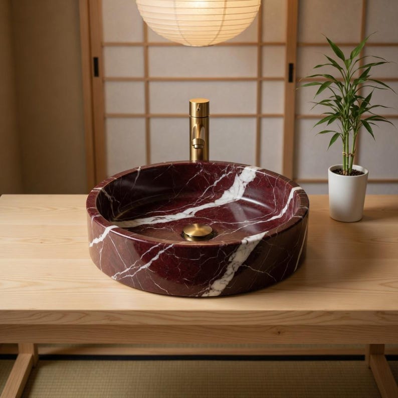 May include: A circular vessel sink crafted from deep red marble with prominent white veining. The basin features a central brass drain plug and is paired with a tall, cylindrical brass faucet fixture centered directly behind the bowl.
