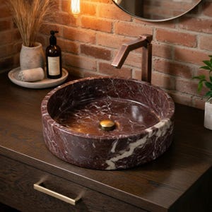 May include: A circular vessel sink crafted from deep red marble with prominent white veining. The sink features a polished finish and a central brass-toned drain cover, paired with a tall, angular, metallic copper-toned faucet fixture.