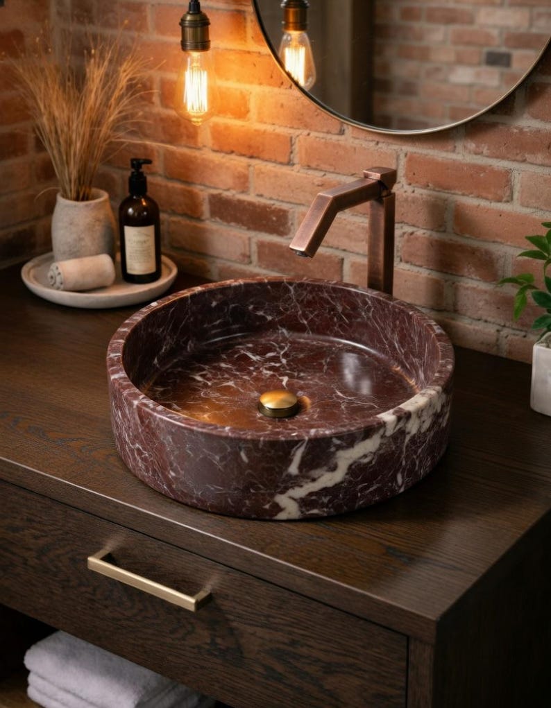 May include: A circular vessel sink crafted from deep red marble with prominent white veining. The sink features a polished finish and a central brass-toned drain cover, paired with a tall, angular, metallic copper-toned faucet fixture.