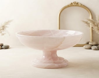 May include: A pale pink pedestal bowl crafted from smooth, polished stone. The bowl features a wide, shallow basin with a rounded rim, supported by a sturdy, circular base. The surface displays natural, subtle veining throughout the material.