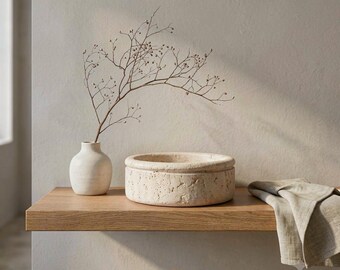 May include: A small, off-white ceramic vase containing a thin, bare branch with tiny buds, positioned next to a round, porous, beige stone bowl with a textured surface and a folded light beige linen cloth draped over the edge of the wooden shelf.