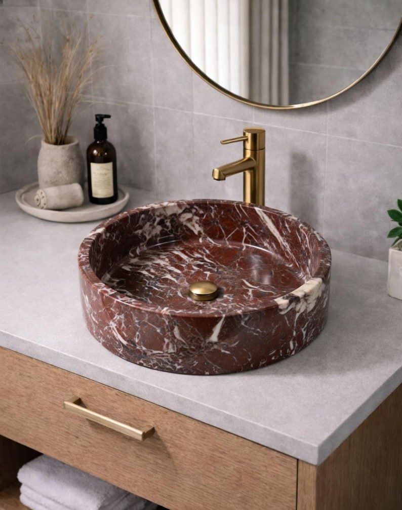 May include: A round vessel sink made of deep red marble with prominent white veining, featuring a polished finish. The sink is equipped with a central gold-toned drain plug and is paired with a matching gold-toned faucet mounted on the countertop.