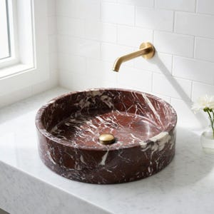 May include: A round vessel sink crafted from deep red marble with prominent white veining. The sink features a central brass drain cover and is paired with a wall-mounted brass faucet, positioned above a white marble countertop surface.