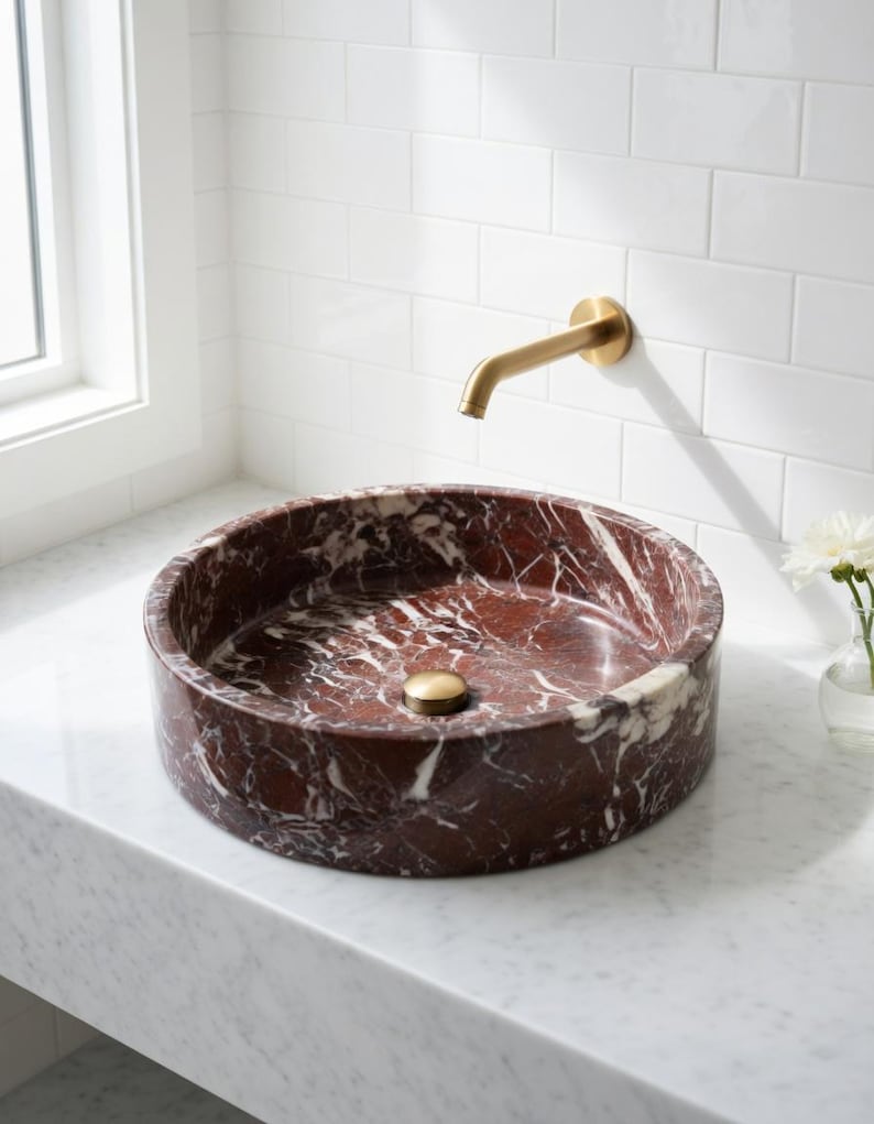 May include: A round vessel sink crafted from deep red marble with prominent white veining. The sink features a central brass drain cover and is paired with a wall-mounted brass faucet, positioned above a white marble countertop surface.